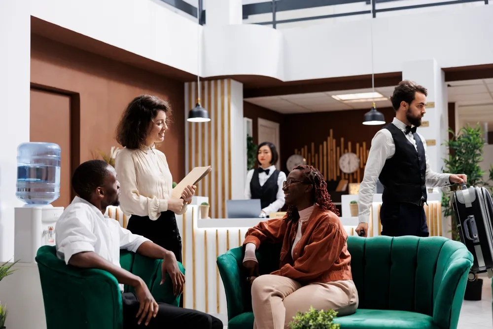 Hotelreceptie waar medewerkers gasten verwelkomen; mensen zitten in de lounge te wachten terwijl een receptionist helpt en een hotelgast met een koffer voorbijloopt.