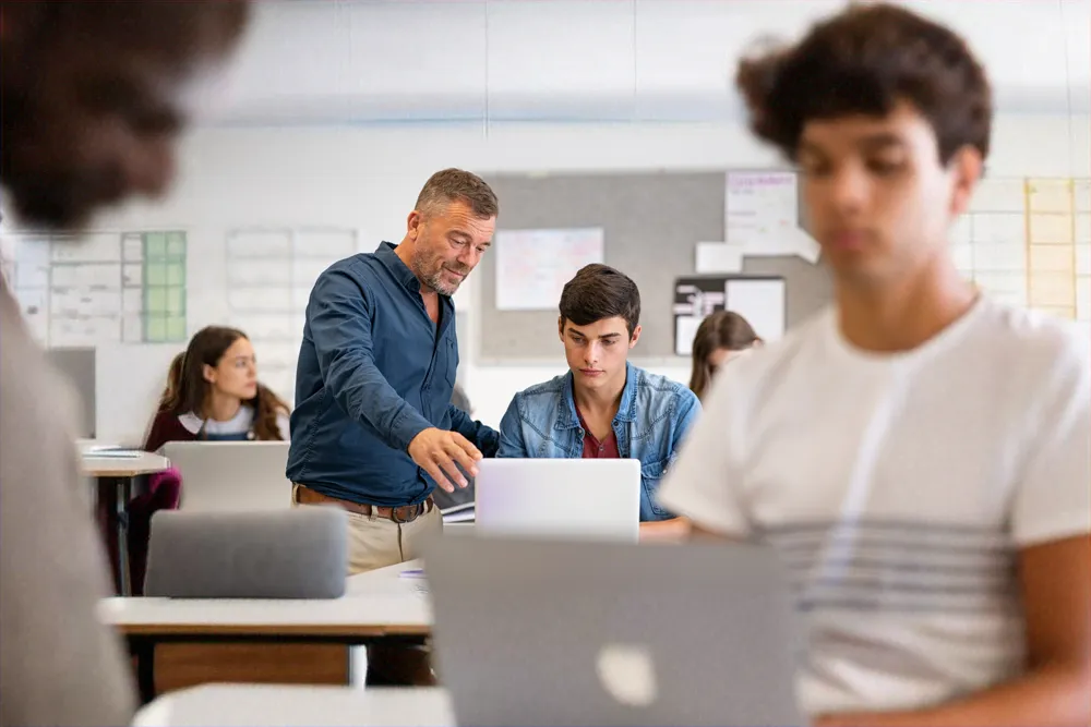 Docent die een student helpt bij zijn laptop in een klaslokaal, terwijl andere studenten aan hun eigen werkstations zitten.
