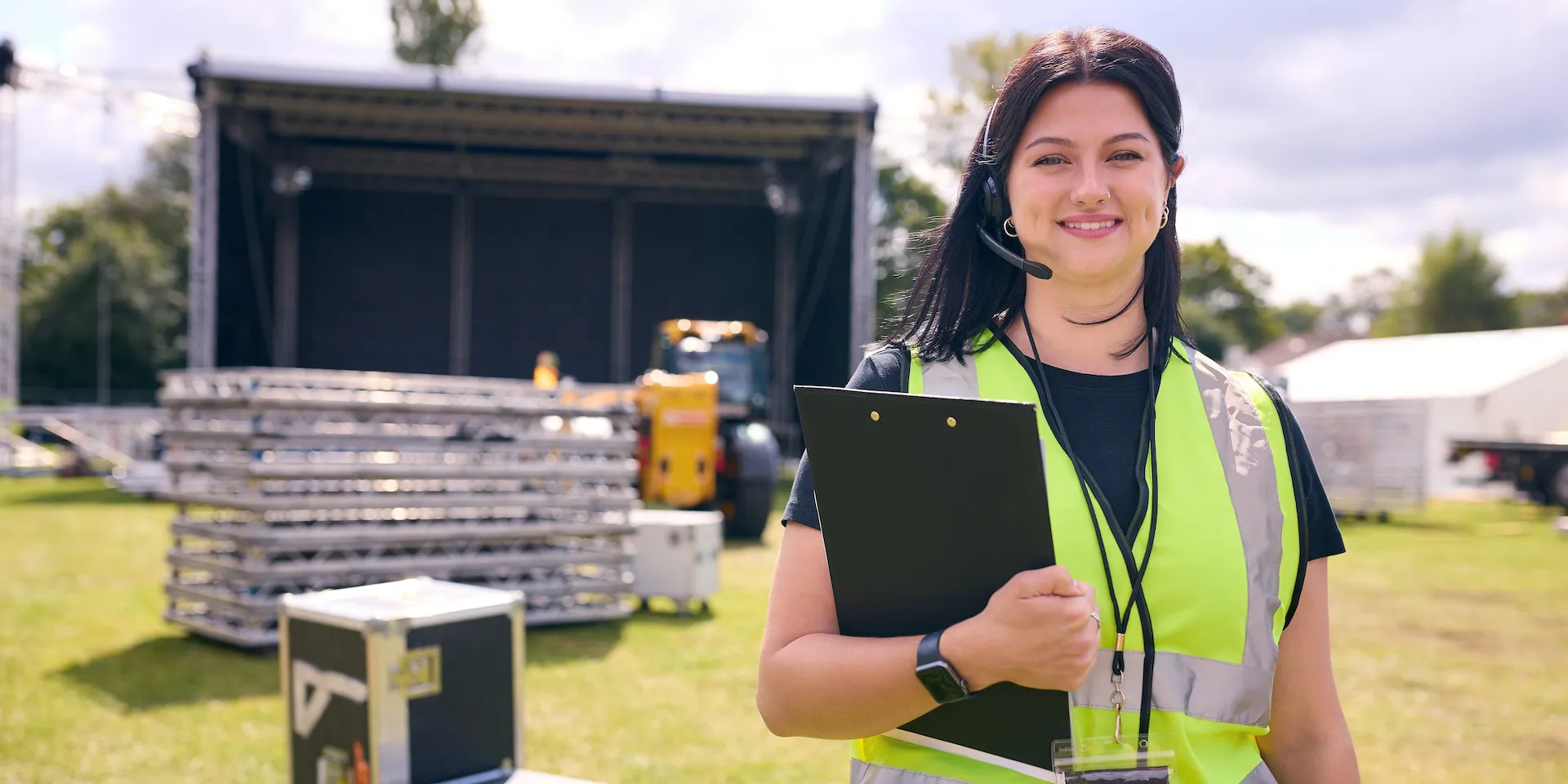 Vrouwelijke evenementmedewerker met een veiligheidshesje houdt een clipboard vast op een buitenlocatie waar een podium en apparatuur worden opgebouwd.