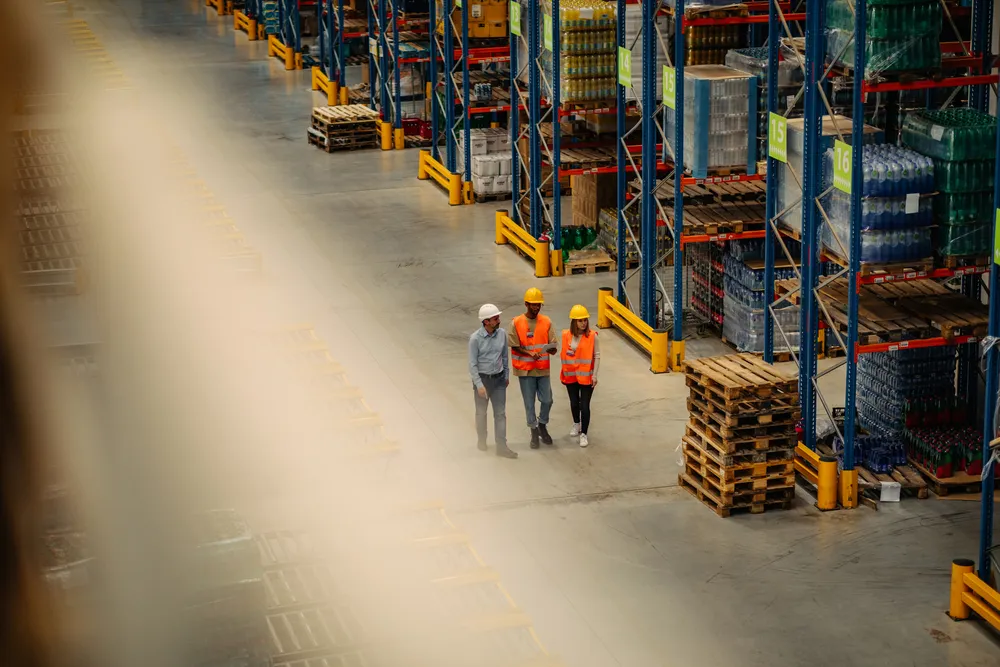 Drie medewerkers met helmen en veiligheidshesjes lopen door een grote opslaghal tussen hoge stellingen met goederen en pallets.