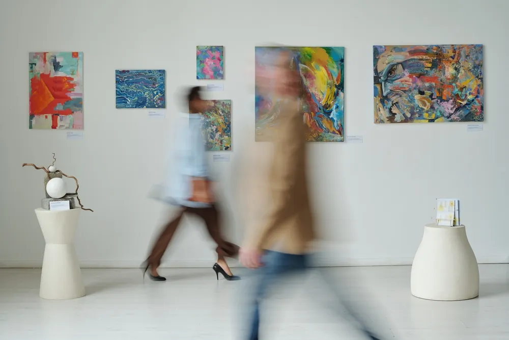 Bezoekers lopen door een kunstgalerie met kleurrijke schilderijen aan de muur en enkele sculpturen op sokkels.