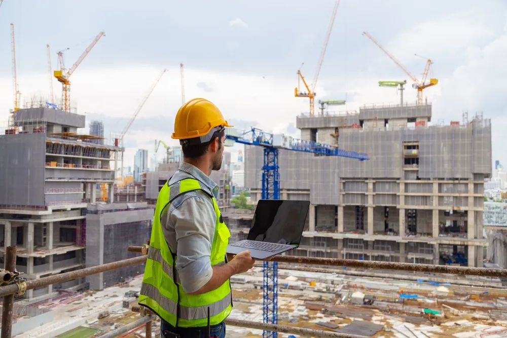 Bouwmedewerker met veiligheidshelm en reflectievest kijkt uit over een grote bouwplaats terwijl hij een laptop vasthoudt.