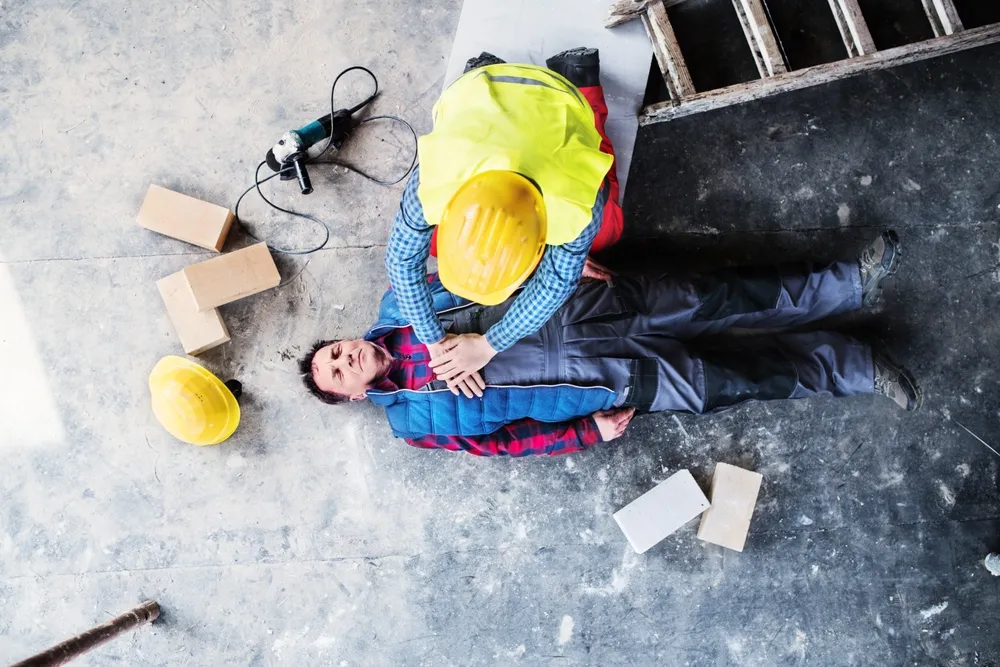 Bouwmedewerker verleent eerste hulp aan een collega die op de grond ligt na een ongeluk op de werkplaats.